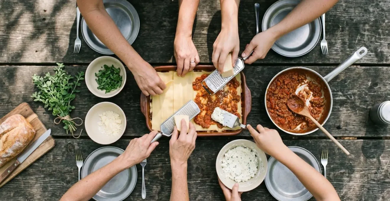 Mani di diverse persone che preparano insieme le lasagne su un tavolo da campeggio all'aperto